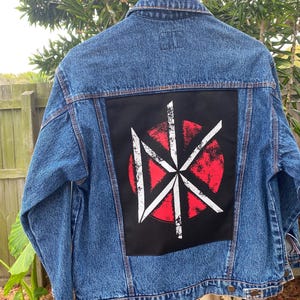 May include: Blue denim jacket with a large black patch on the back featuring a white and red graphic design. The jacket has a classic design with button closures and a collared neckline. The jacket is hanging on a wooden hanger.