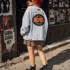 May include: A light blue denim jacket featuring a large circular AC/DC patch on the back with red and yellow lettering, a smaller red AC/DC logo near the collar, a floral print skirt, and black leather lace-up boots with yellow pull tabs.