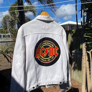 May include: A white denim jacket featuring a large circular AC/DC logo on the back. The black circular patch displays the red and yellow AC/DC band logo with the text High Voltage Rock N' Roll, Australia, and Est. 1973 printed in yellow.