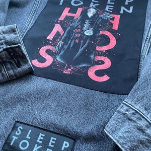 May include: A grey denim jacket featuring a large rectangular back patch with a graphic of a hooded figure and the text SLEEP TOKEN. The patch includes red and white accents. A smaller rectangular patch on the lower section reads SLEEP TOKEN.