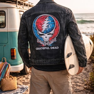 May include: Black denim jacket with a colorful Grateful Dead logo on the back. The logo features a skull, lightning bolt, and text that reads "Grateful Dead". The person is holding a surfboard on a sandy beach.