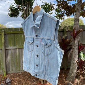May include: A light blue denim vest featuring a classic collar, a front button closure with dark metal buttons, and two chest pockets with buttoned flaps. A small red tab is visible on the left chest pocket. The vest is sleeveless with raw edges.