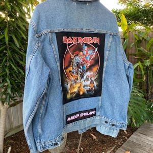 Puede incluir: Una chaqueta de mezclilla azul claro con un parche rectangular grande en la espalda que presenta un gráfico de Iron Maiden de una figura a caballo sosteniendo una bandera Union Jack. Debajo hay un parche con el texto Maiden England.