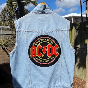 Puede incluir: Un chaleco de mezclilla azul claro con un gran parche circular en la parte posterior. El parche es negro con el logotipo de AC/DC en rojo y amarillo, incluyendo el texto HIGH VOLTAGE ROCK N' ROLL, AUSTRALIA y EST. 1973. Tiene cuello y botones.