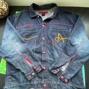 May include: A dark blue denim jacket featuring red contrast stitching, a button-down front, and long sleeves with buttoned cuffs. The chest displays the embroidered text AVIREX in red and a large yellow letter A. It includes two chest pockets.
