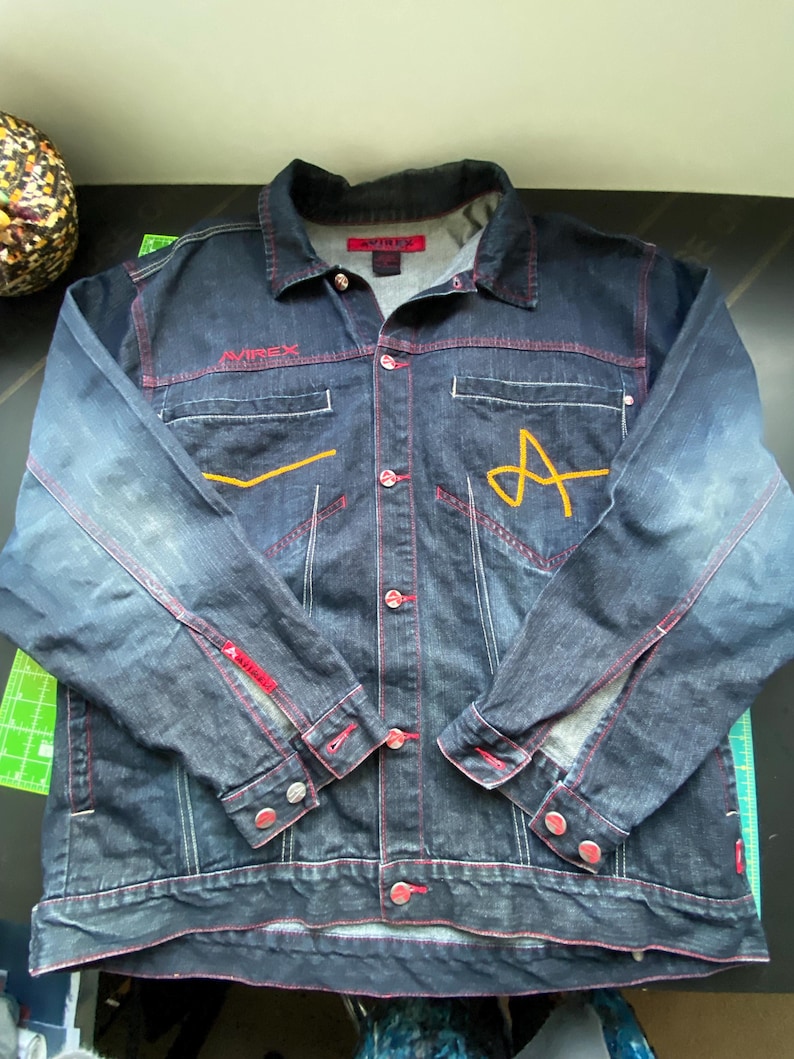 May include: A dark blue denim jacket featuring red contrast stitching, a button-down front, and long sleeves with buttoned cuffs. The chest displays the embroidered text AVIREX in red and a large yellow letter A. It includes two chest pockets.