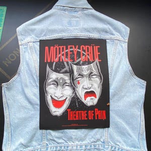 May include: A light blue acid-wash denim vest featuring a large rectangular black patch on the back. The patch displays the text Mötley Crüe and Theatre of Pain in red, with a graphic of two theatrical masks, one smiling and one crying.