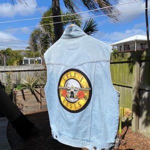 May include: A light blue denim vest featuring a large circular Guns N' Roses logo on the back. The graphic displays two crossed revolvers, two red roses, and the band name in black text on a yellow background. The vest has a collar and metal buttons.