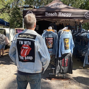 May include: A light blue denim jacket featuring a large rectangular patch on the back with the Rolling Stones logo, the text I'M WITH THE BAND, and the band name. The jacket is displayed among other denim jackets with various band patches.