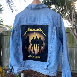 May include: A light blue denim jacket featuring a large rectangular black patch on the back. The patch displays the yellow Metallica logo above a silhouette of four figures against a bright light, with the text SEVENTY-TWO printed in yellow below.