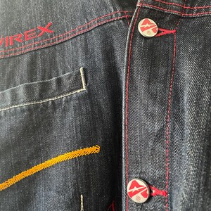 May include: A dark blue denim jacket featuring red contrast stitching and the text VIREX embroidered in red. The jacket includes silver-toned metal buttons with a red and white logo design and a decorative yellow zigzag stitch pattern on the front.