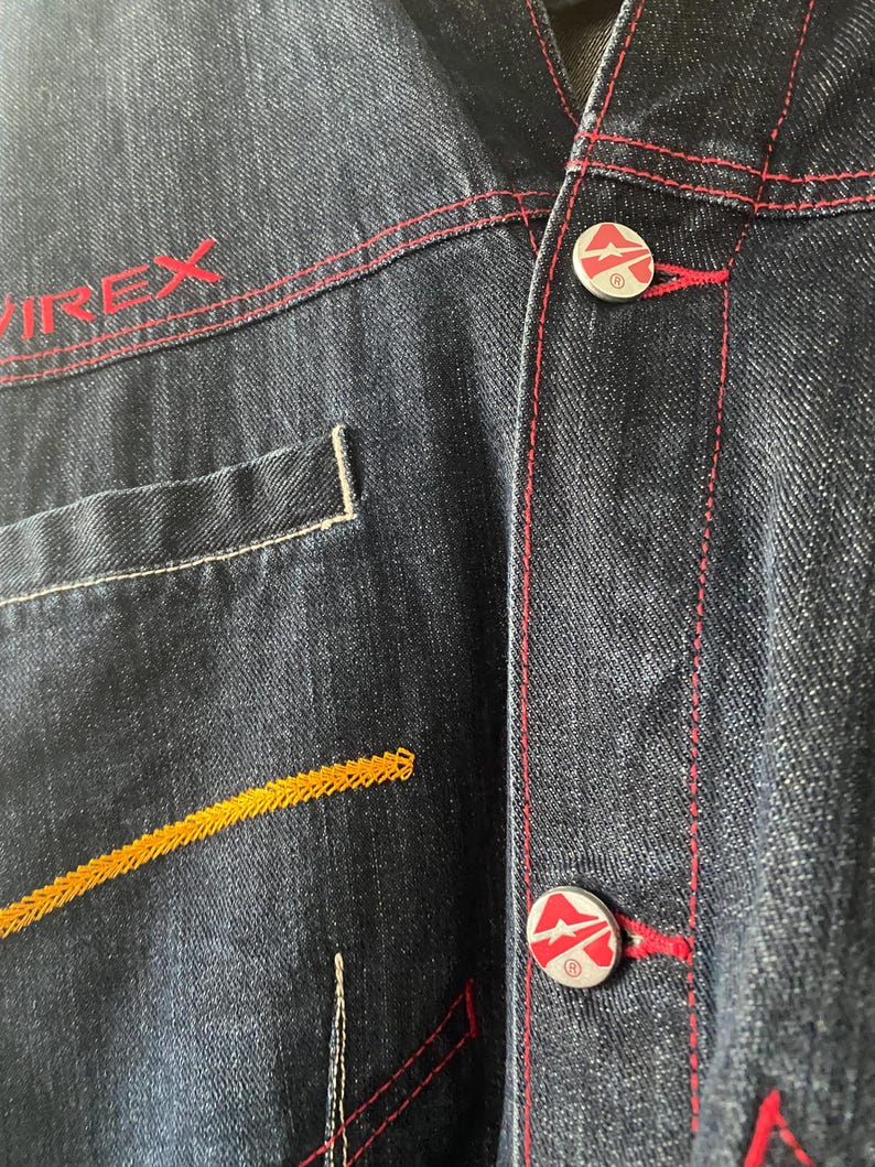May include: A dark blue denim jacket featuring red contrast stitching and the text VIREX embroidered in red. The jacket includes silver-toned metal buttons with a red and white logo design and a decorative yellow zigzag stitch pattern on the front.
