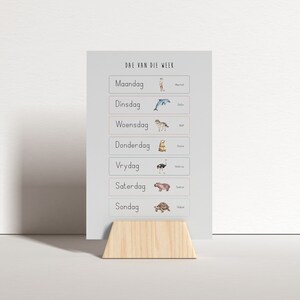 Könnte beinhalten: Ein weißes rechteckiges Schild mit dem Text "Dae van die Week" und den Wochentagen auf Afrikaans, jeweils mit einer Tierillustration. Das Schild wird von einem hellen Holzständer getragen.