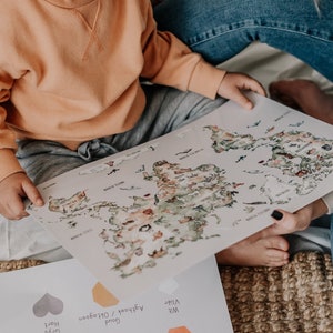 Op de afbeelding: Een kind en een volwassene houden een wereldkaartprint vast met kleurrijke illustraties van continenten en oceanen. De kaart is lichtgekleurd met een aquarel-stijl. Een tweede vel papier met kleurstalen is zichtbaar.