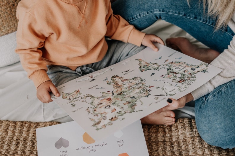 Op de afbeelding: Een kind en een volwassene houden een wereldkaartprint vast met kleurrijke illustraties van continenten en oceanen. De kaart is lichtgekleurd met een aquarel-stijl. Een tweede vel papier met kleurstalen is zichtbaar.