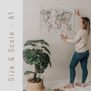 Op de afbeelding: Een grote A1-formaat wereldkaart poster met kleurrijke illustraties van dieren en bezienswaardigheden. De kaart wordt tegen een witte muur gehouden. De tekst "Size & Scale - A1" staat aan de linkerkant van de afbeelding. Ongeveer 59 x 84 cm.