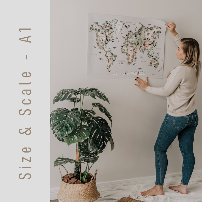 Op de afbeelding: Een grote A1-formaat wereldkaart poster met kleurrijke illustraties van dieren en bezienswaardigheden. De kaart wordt tegen een witte muur gehouden. De tekst "Size & Scale - A1" staat aan de linkerkant van de afbeelding. Ongeveer 59 x 84 cm.