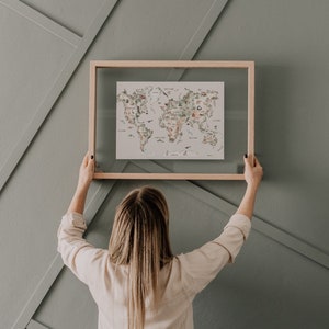 Op de afbeelding: Een wereldkaartprint met een lichte houten lijst. De kaart bevat kleurrijke illustraties van bezienswaardigheden en dieren. De print wordt tegen een grijze muur met geometrische panelen gehouden.
