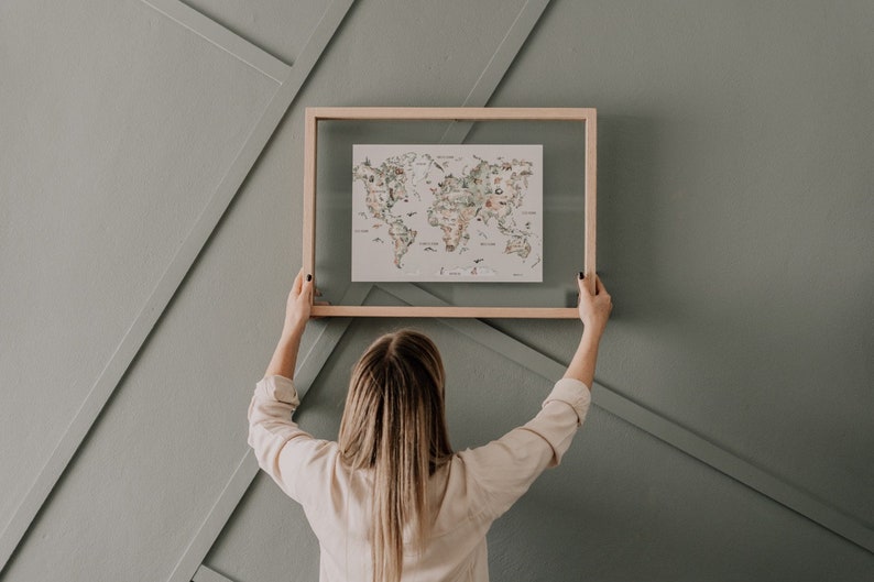 Op de afbeelding: Een wereldkaartprint met een lichte houten lijst. De kaart bevat kleurrijke illustraties van bezienswaardigheden en dieren. De print wordt tegen een grijze muur met geometrische panelen gehouden.