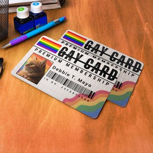 Op de afbeelding: Twee "GAY CARD" lidmaatschapskaarten met regenboogaccenten en een foto van een kat. De kaarten hebben de tekst "PREMIUM MEMBERSHIP" en de naam "Debbie T. Mayo". Een pen, inktflessen en een bril zijn ook zichtbaar.