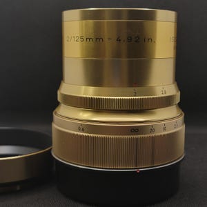 Op de afbeelding: Een goudkleurige cameralens met zwarte accenten. De lens heeft de tekst "2/125 mm - 4,92 in." en diverse meetmarkeringen. Een zonnekap ligt links.