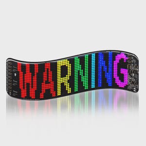Può includere: Un display LED nero curvo con la scritta "WARNING" in colori arcobaleno. Il display utilizza piccole luci colorate per formare le lettere. Lo sfondo è grigio chiaro.