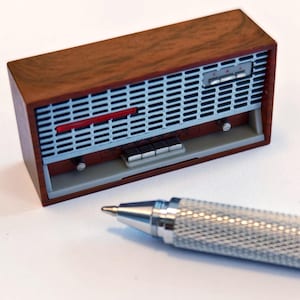 Könnte beinhalten: Ein Miniatur-Vintage-Radio mit einem braunen Holzgehäuse und einem silbernen Lautsprechergrill. Das Radio hat einen roten Akzentstreifen und silberne Knöpfe. Ein silberner Stift mit strukturierter Oberfläche befindet sich im Vordergrund.