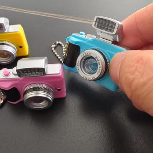 May include: Miniature toy cameras in blue, pink, and yellow. Each camera has a silver lens, flash, and a black grip. The blue camera has the text "Zhaoxiangji Optical" and lens specifications. These small cameras are likely keychains or decorative items.