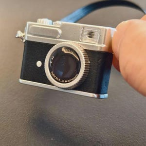 May include: A miniature camera charm with a silver top and black body. The camera has a prominent lens with text and a blue strap. The charm is held by a hand against a dark background. The camera is a vintage style.
