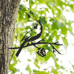 Könnte beinhalten: Schwarze Metallsilhouette eines Kardinals, der auf einem Ast mit einer Kiefernzapfen sitzt. Der Kardinal ist nach links gerichtet und hat einen langen Schwanz. Der Ast ist kahl und hat ein paar kleine Blätter.