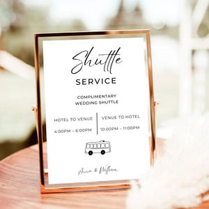 May include: A framed sign with the words "Shuttle Service" in elegant script. The sign details a complimentary wedding shuttle service, including times for hotel to venue and venue to hotel. The names "Anna & William" are at the bottom.