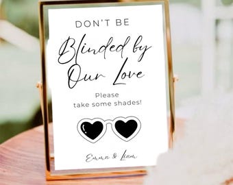 May include: A framed sign with the text "DON'T BE Blinded by Our Love" and "Please take some shades!" in black script. Below the text is a graphic of heart-shaped sunglasses. The frame is gold.