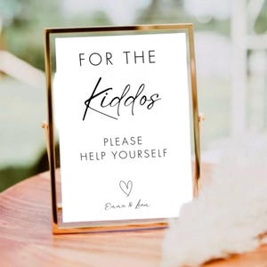 Puede incluir: Un letrero enmarcado con borde dorado y fondo blanco. El letrero dice "FOR THE Kiddos PLEASE HELP YOURSELF" en escritura negra. Un pequeño corazón y los nombres "Emma & Liam" están en la parte inferior.