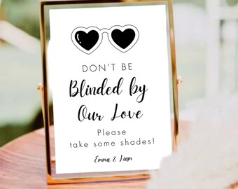 May include: A framed sign with a gold border. The sign reads "DON'T BE Blinded by Our Love" with heart-shaped sunglasses above the text. Below the text, it says "Please take some shades!" and the names "Emma & Liam".