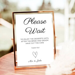 May include: A gold-colored framed sign with the words "Please Wait" in elegant script. The sign includes the text "TO ENJOY THE DESSERTS UNTIL AFTER THE BRIDE AND GROOM HAVE CUT THE CAKE" and "THANK YOU!" with a heart and names "Mia & Jake".