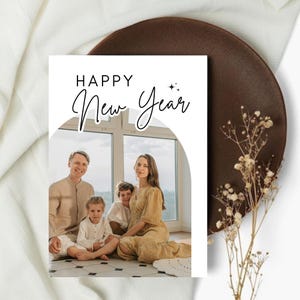 Puede incluir: Una tarjeta de felicitación blanca con las palabras "HAPPY New Year" en escritura negra. La tarjeta presenta un retrato familiar dentro de un arco blanco. El fondo incluye un plato marrón y flores secas.