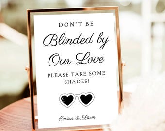 May include: A framed sign with the text "DON'T BE Blinded by Our Love PLEASE TAKE SOME SHADES!" in black script. Below the text is a graphic of heart-shaped sunglasses. The sign is in a gold frame and includes the names "Emma & Liam".