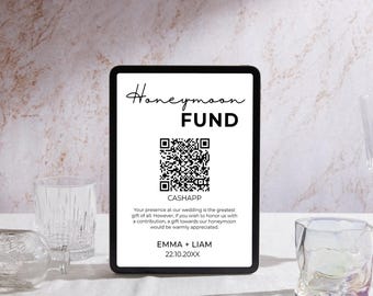 May include: A digital display with the words "Honeymoon FUND" in black script. Below is a QR code and the text "CASHAPP". The display also includes a message about wedding gifts and the names "EMMA + LIAM" with a date. The display is on a white surface.