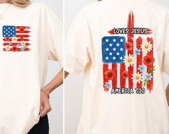 May include: Cream-colored t-shirt featuring an American flag design with red stripes, blue stars, and floral accents. The back of the shirt has the text "LOVES JESUS AMERICA TOO" with a cross and flag design.