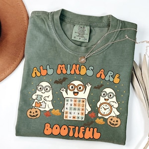 Puede incluir: Camiseta verde Comfort Colors con el texto "ALL MINDS ARE BOOTIFUL" en naranja y azul. La camiseta presenta tres fantasmas de dibujos animados con gafas, sosteniendo una pieza de rompecabezas, un tablero de alfabeto y un reloj. También se muestran calabazas y murciélagos.