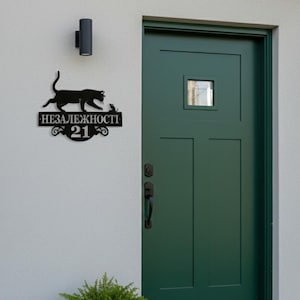 May include: Black metal house number sign featuring a cat silhouette, the text "НЕЗАЛЕЖНОСТІ 21", and decorative elements. The sign is mounted on a white exterior wall near a green door and a potted plant.