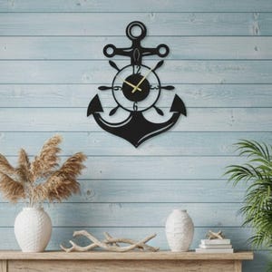 Könnte beinhalten: Eine schwarze Metall-Anker-Wanduhr mit rundem Zifferblatt und goldenen Zeigern. Die Uhr ist an einer hellblauen Holzwand befestigt. Das Design der Uhr umfasst ein Steuerrad und einen Anker, die ein maritimes Thema hervorrufen.
