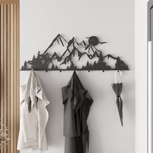 May include: A black metal wall-mounted coat rack featuring a mountain range silhouette with trees and a sun. The rack has multiple hooks for hanging coats, towels, or umbrellas. The items hanging on the rack include a light-colored towel, a dark gray robe, and a black umbrella.