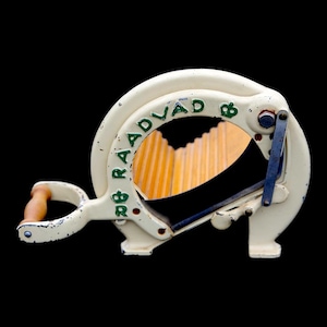 May include: Off-white vintage bread slicer with the text "RAADVAD" in green. The slicer has a wooden handle and a blue lever. The blade is visible through the circular opening. The background is black.