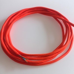 May include: A coil of bright orange electrical cable. The cable is covered in a woven fabric and has exposed internal wires showing blue, brown, and copper colours. This type of cable is commonly used for lighting and other electrical applications.