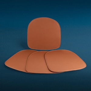 Op de afbeelding: Vier bruine leren stoelkussens tegen een donkerblauwe achtergrond. De kussens, in een warme, aardse tint, hebben afgeronde randen en een gladde textuur. Eén kussen is boven de andere geplaatst.