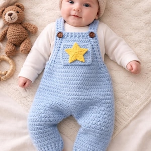 Puede incluir: Un bebé con un mono de ganchillo azul claro con una estrella amarilla en el pecho. El mono tiene botones marrones y se combina con un gorro de punto beige. Un pequeño osito de peluche y un juguete de madera también son visibles.