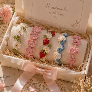 May include: A presentation box with five handmade crochet bracelets. The bracelets feature daisy, heart, strawberry, seashell, and bow designs in white, pink, and blue. The box includes a card that says "Handmade with love."