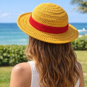 Può includere: Un cappello giallo all'uncinetto con una fascia rossa. Il cappello è indossato da una persona con lunghi capelli castani ondulati. Lo sfondo presenta un mare e un cielo blu, con fogliame verde.