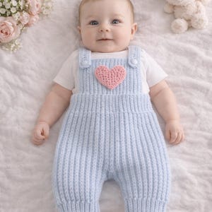 Puede incluir: Un bebé con un pelele de punto azul claro con un corazón rosa. El bebé está acostado sobre una superficie blanca, con un conejito de peluche y flores cerca. La palabra "Lifestyle" está en la parte superior de la imagen.